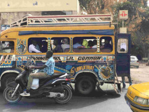A yellow mini bus taxi with writing and drawings on the sid, is parked on a road with a man on a motorbike driving next to it